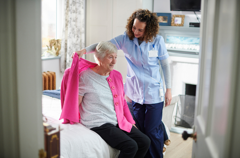 Respite Stay Caregiver helping a senior woman put on a jacket in a warmly furnished apartment at Covina Court Senior Living